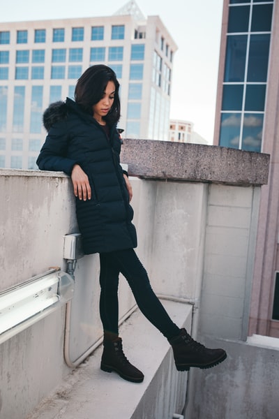 女人穿着黑色大衣外套,靠在灰色的混凝土墙
